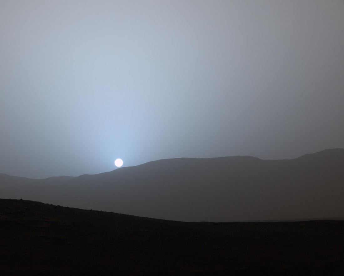 A small Sun setting over the rim of Gale Crater on Mars, surrounded by a soft blue glow